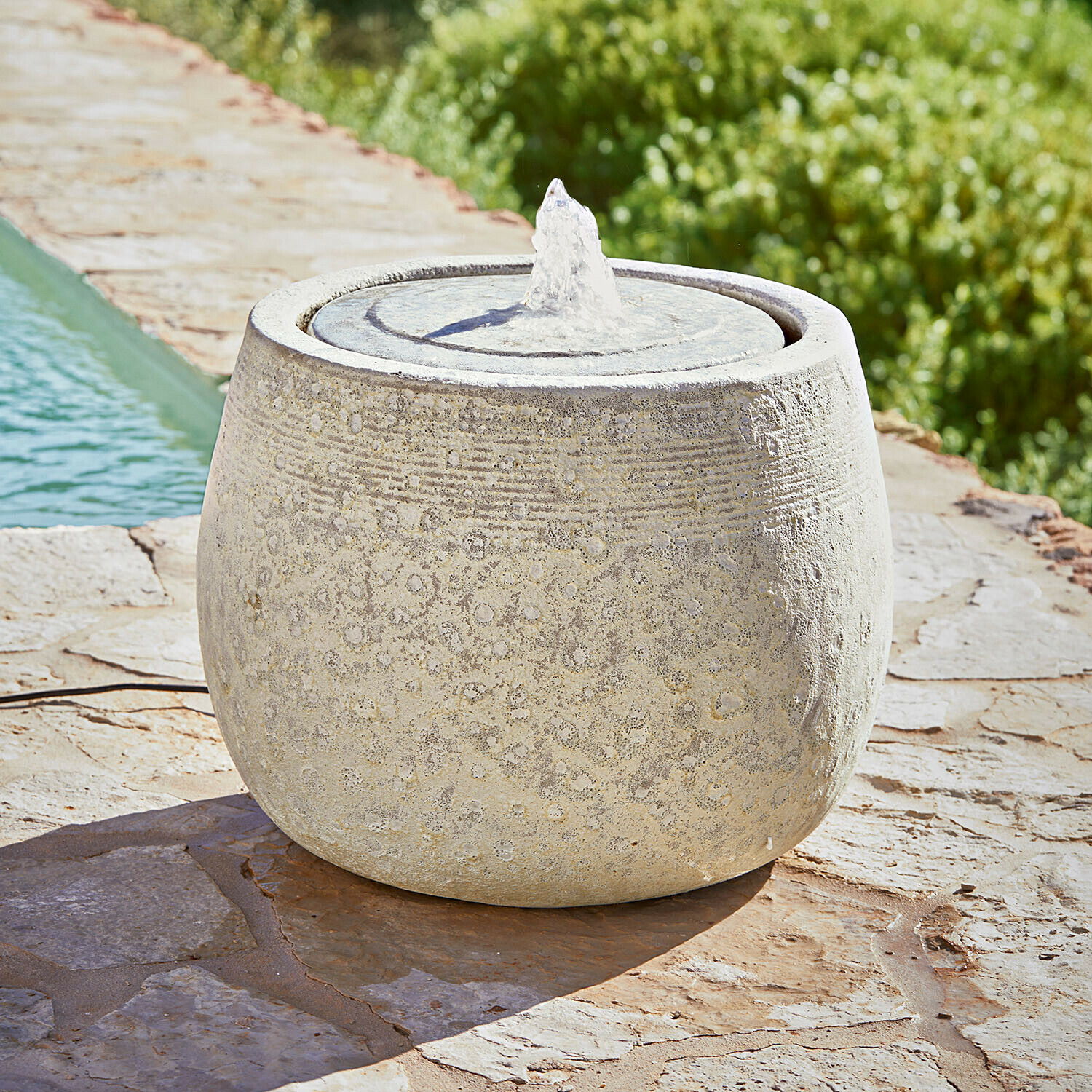 Fontein Seravia Ronde tuinfontein van lichte steen met subtiel patroon en waterstraal op stenen terras