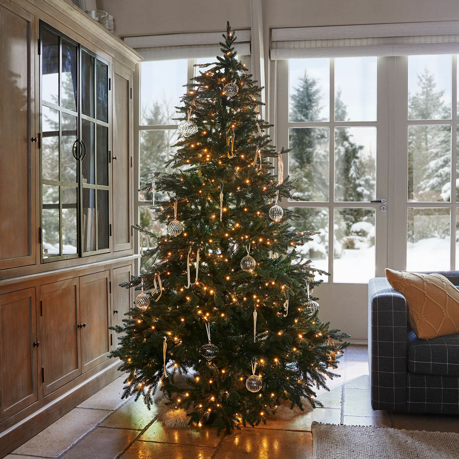 Verlichte kerstboom met groene takken en glazen versieringen in gezellige woonkamer met uitzicht op sneeuw