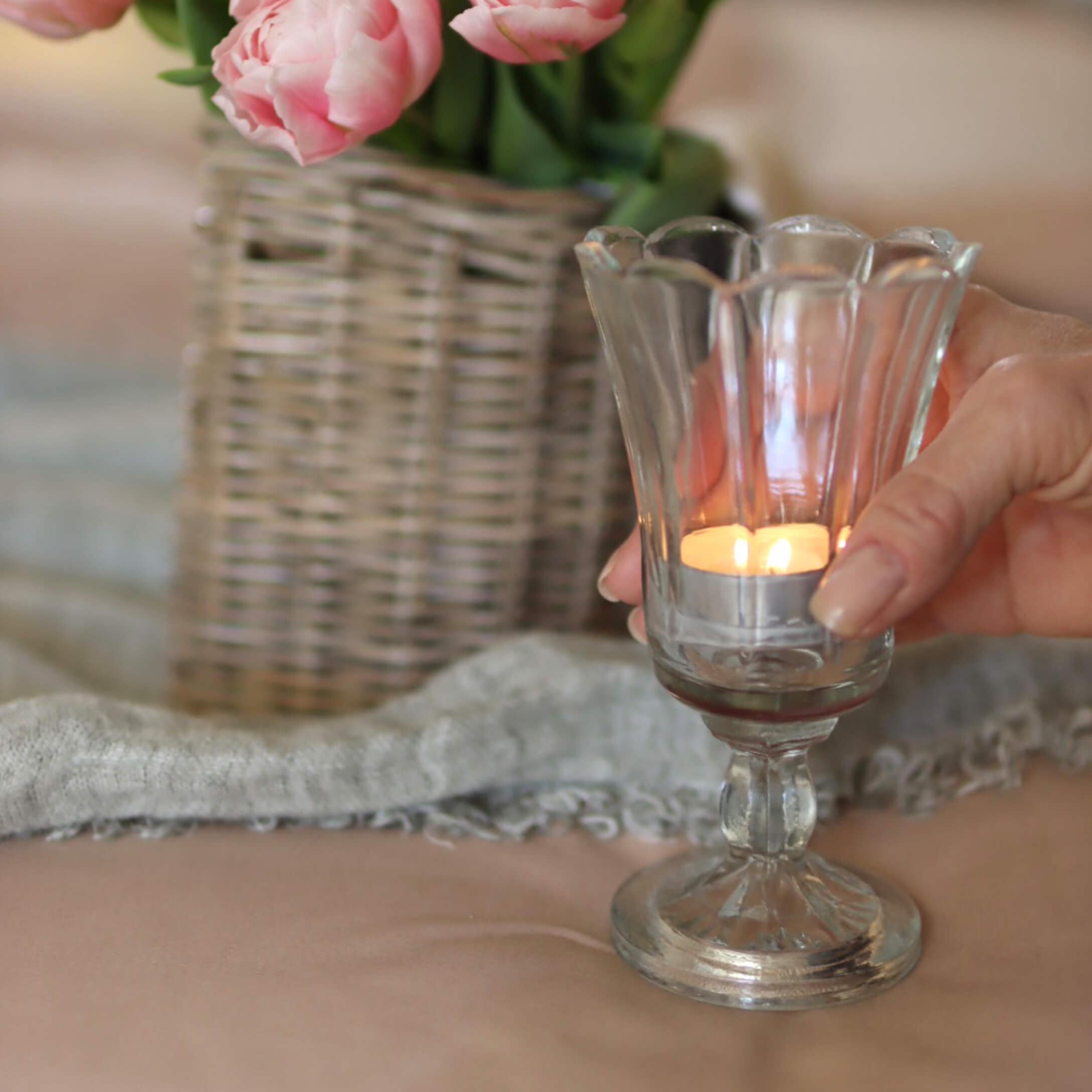 Glazen theelichthouder op voet met brandend kaarsje, vaas met roze bloemen en rieten mand op de achtergrond