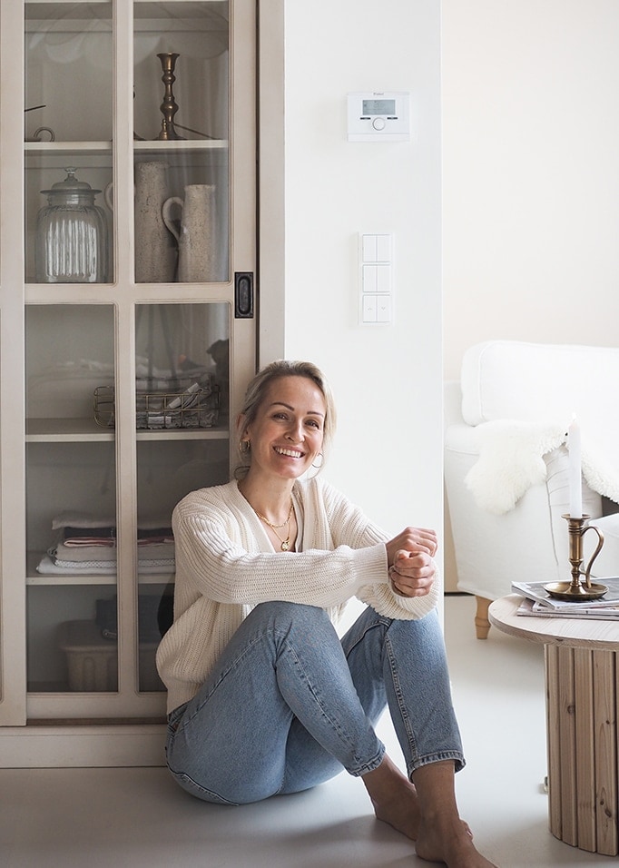 Lachende vrouw in witte trui en blauwe jeans zittend op de vloer in een lichte, gezellige ruimte