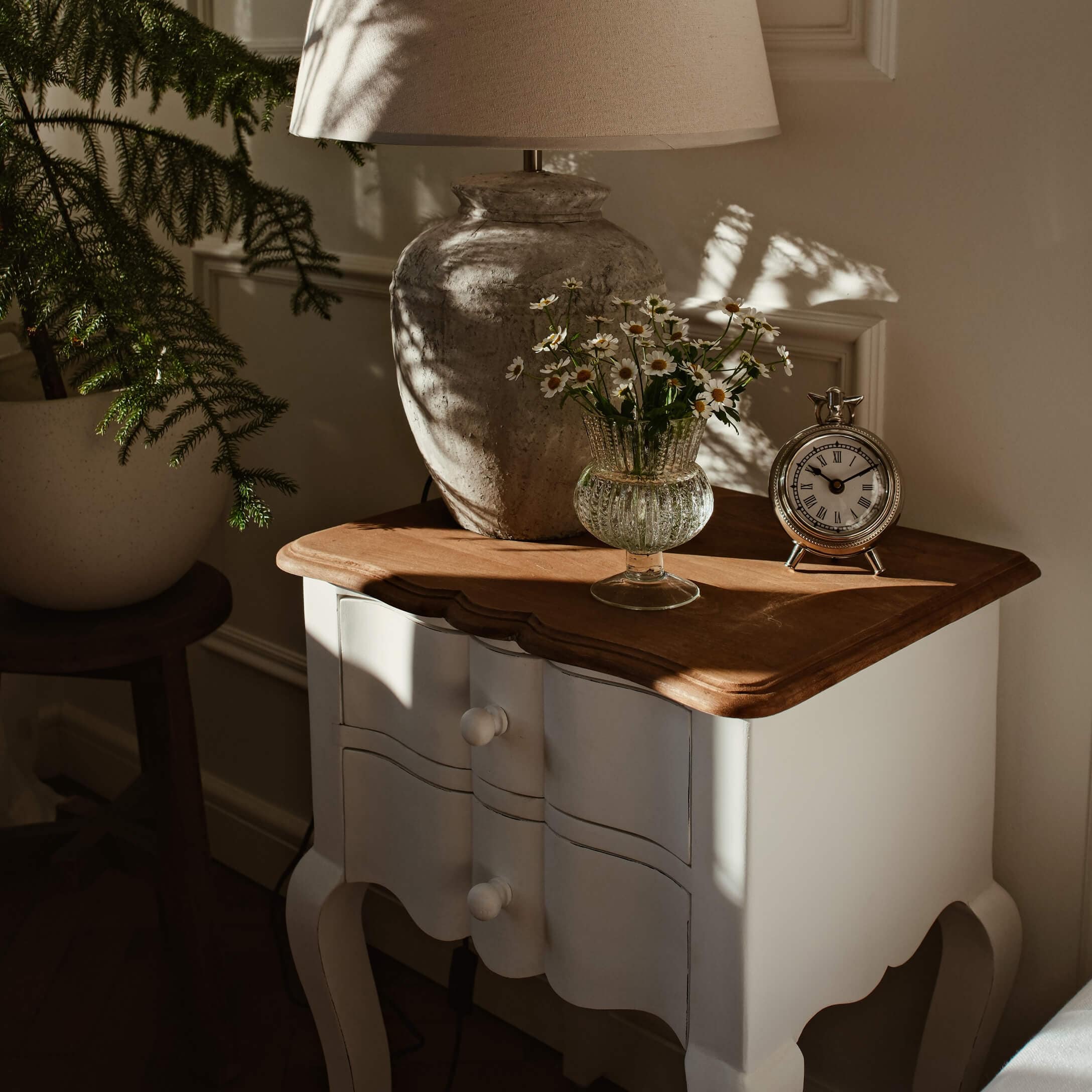Witte houten nachtkast met bruin blad, lamp, glazen vaas met bloemen en zilveren klok in zonlicht