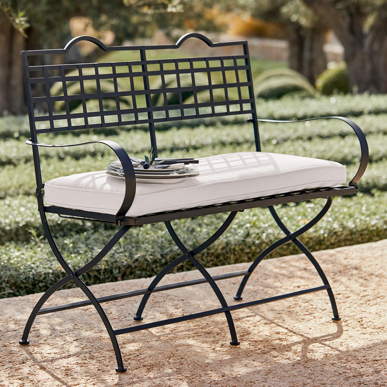 Bank Terell Zwarte metalen tuinbank met wit kussen op stenen terras in de zon