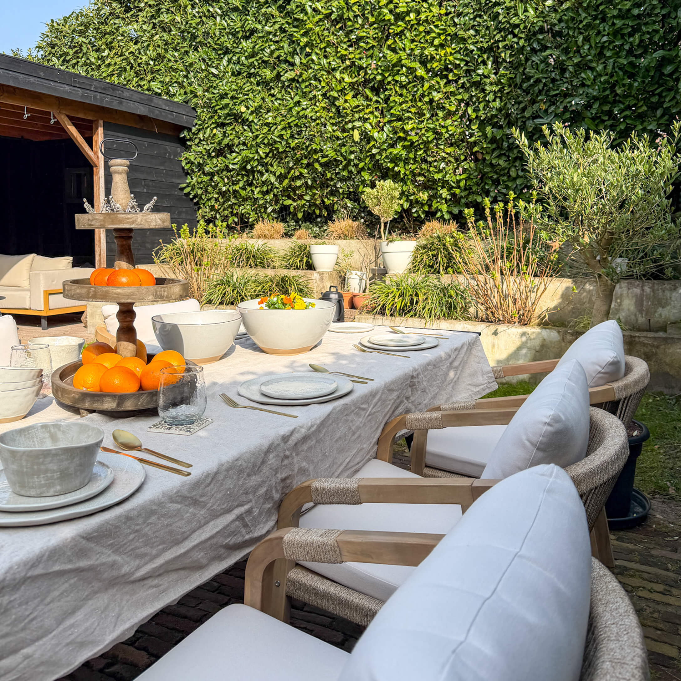 Tuin tafel met wit tafelkleed, keramisch servies en houten fruitstandaard in zonnige tuin