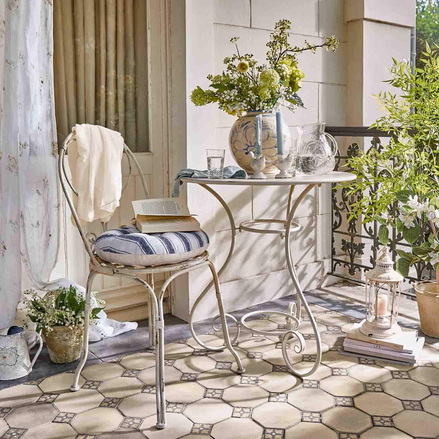 Witte metalen tafel en stoel met gestreept kussen op terras met bloemen en decoratieve lantaarn