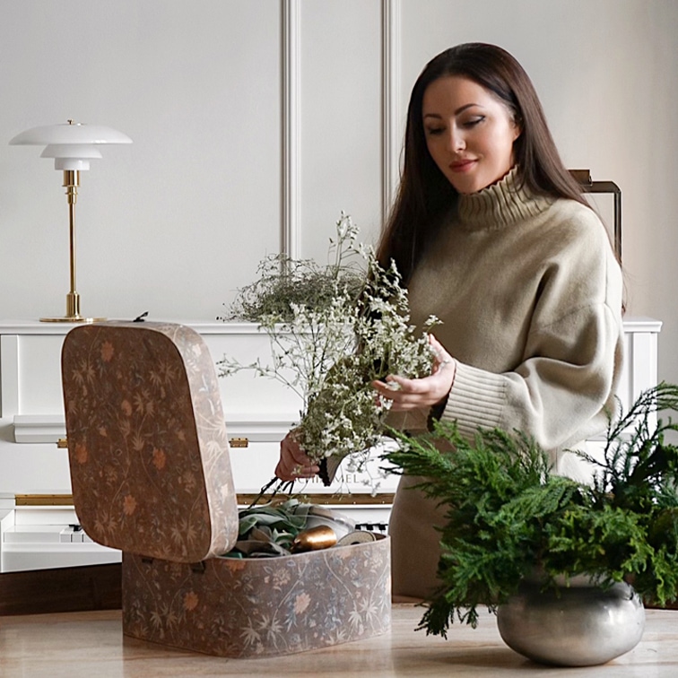 Vrouw in beige trui houdt groene takken vast naast decoratieve doos en plant in zilveren pot