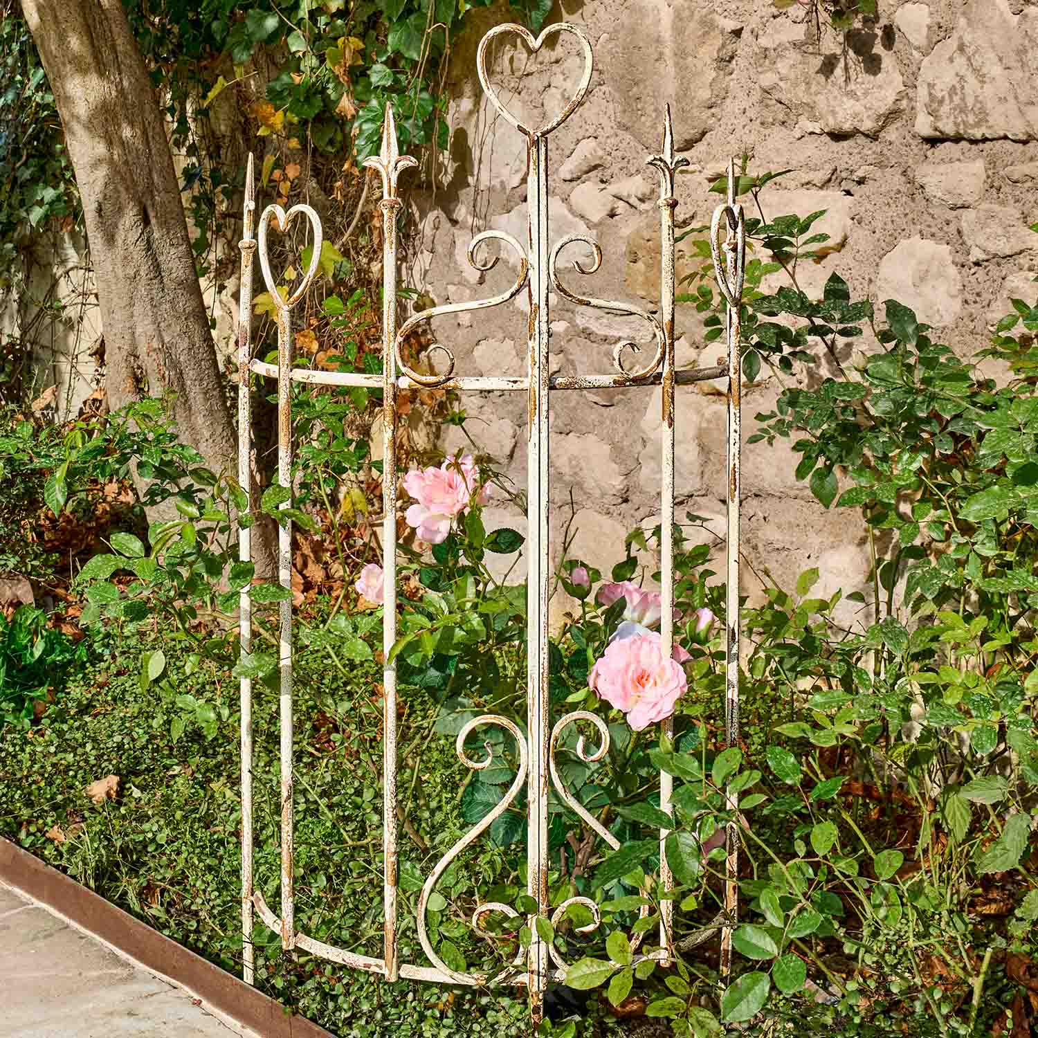 Witte metalen tuinomheining met hartvormige details en roze rozen bij stenen muur in tuin