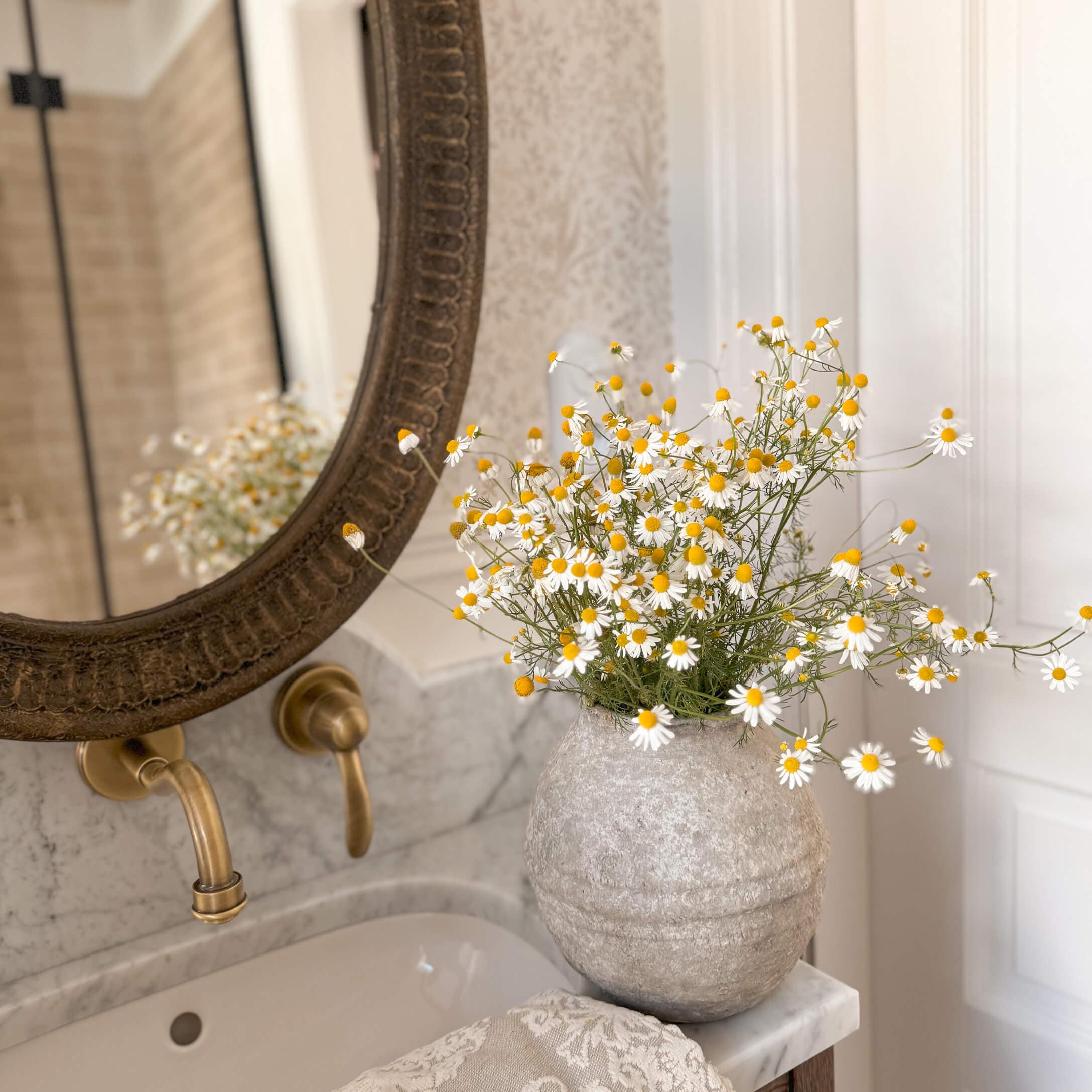 Grijze stenen vaas met witte en gele bloemen op een marmeren wastafel in een stijlvolle badkamer