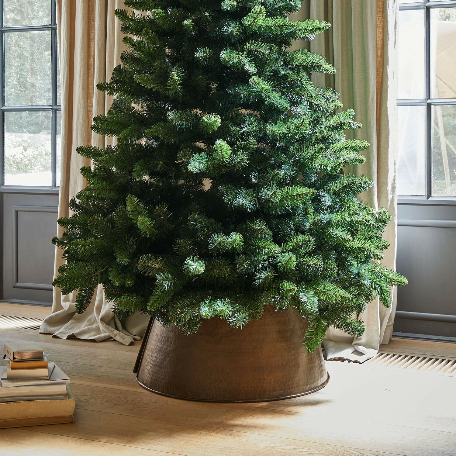 Kerstboomrok Omeyra Kunstkerstboom in koperen metalen pot op lichte houten vloer naast stapel boeken