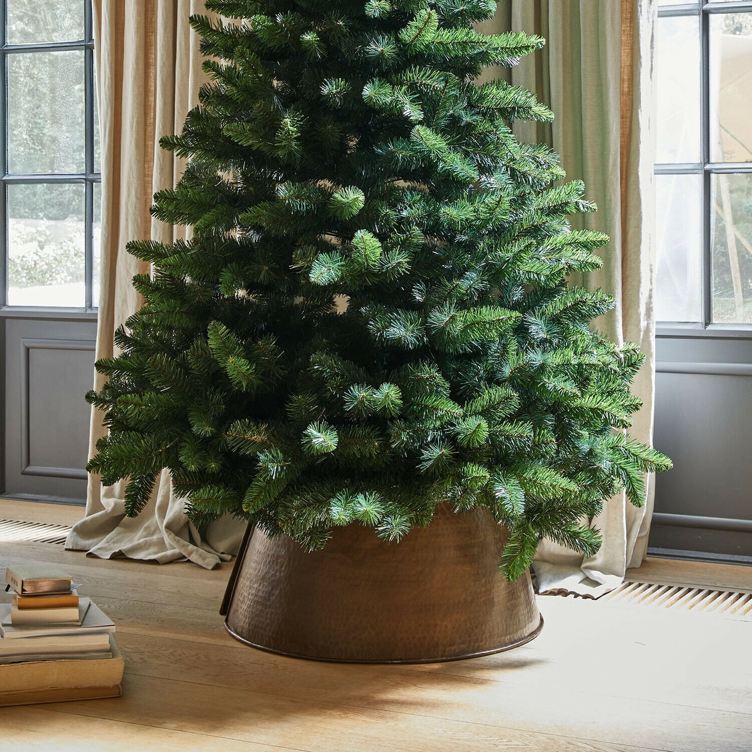Kerstboomrok Omeyra Kunstkerstboom in koperen metalen pot op lichte houten vloer naast stapel boeken