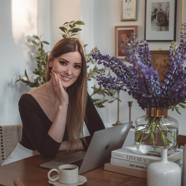 Vrouw aan houten tafel met laptop en glazen vaas met paarse bloemen in lichte kamer
