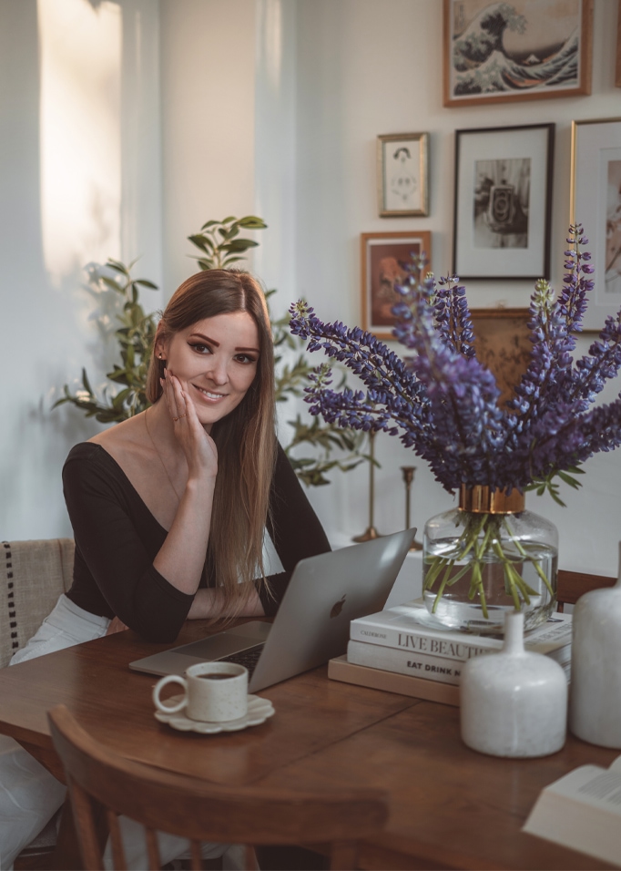 Vrouw aan houten tafel met laptop en glazen vaas met paarse bloemen in lichte kamer