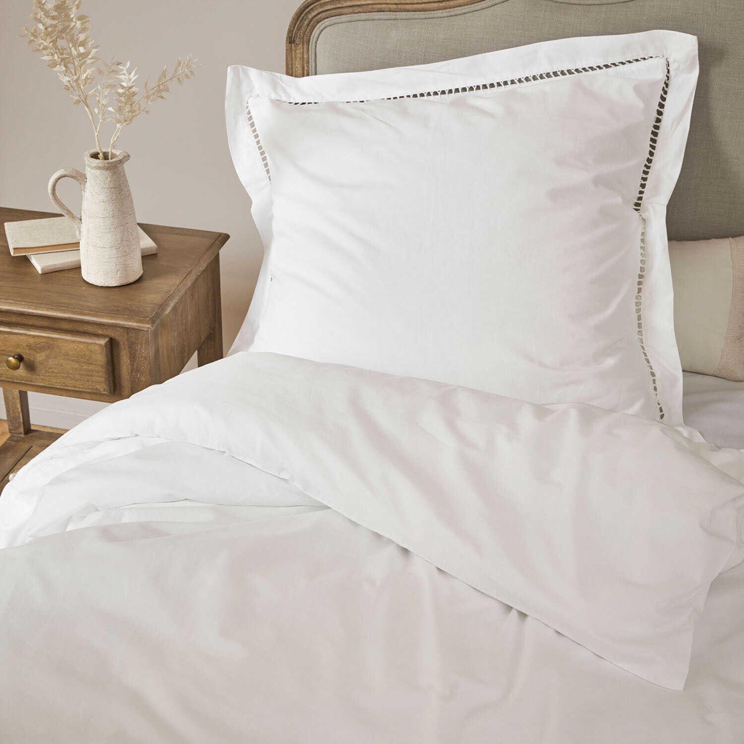 Beddengoed Belvero Witte katoenen kussensloop met decoratieve opengewerkte rand op bed naast houten nachtkastje