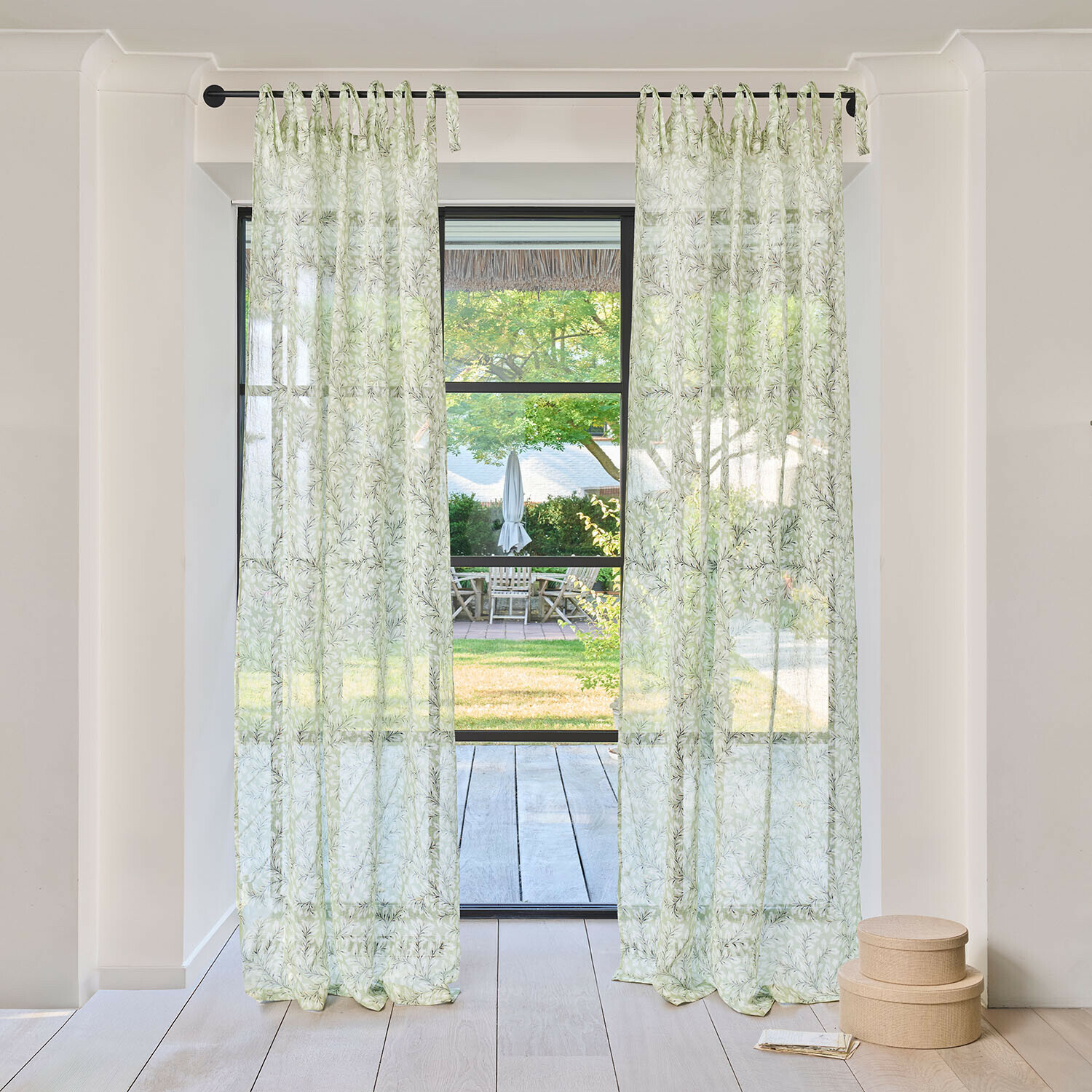 Transparante gordijnen met groen bladmotief aan zwarte roede in lichte kamer met uitzicht op tuin