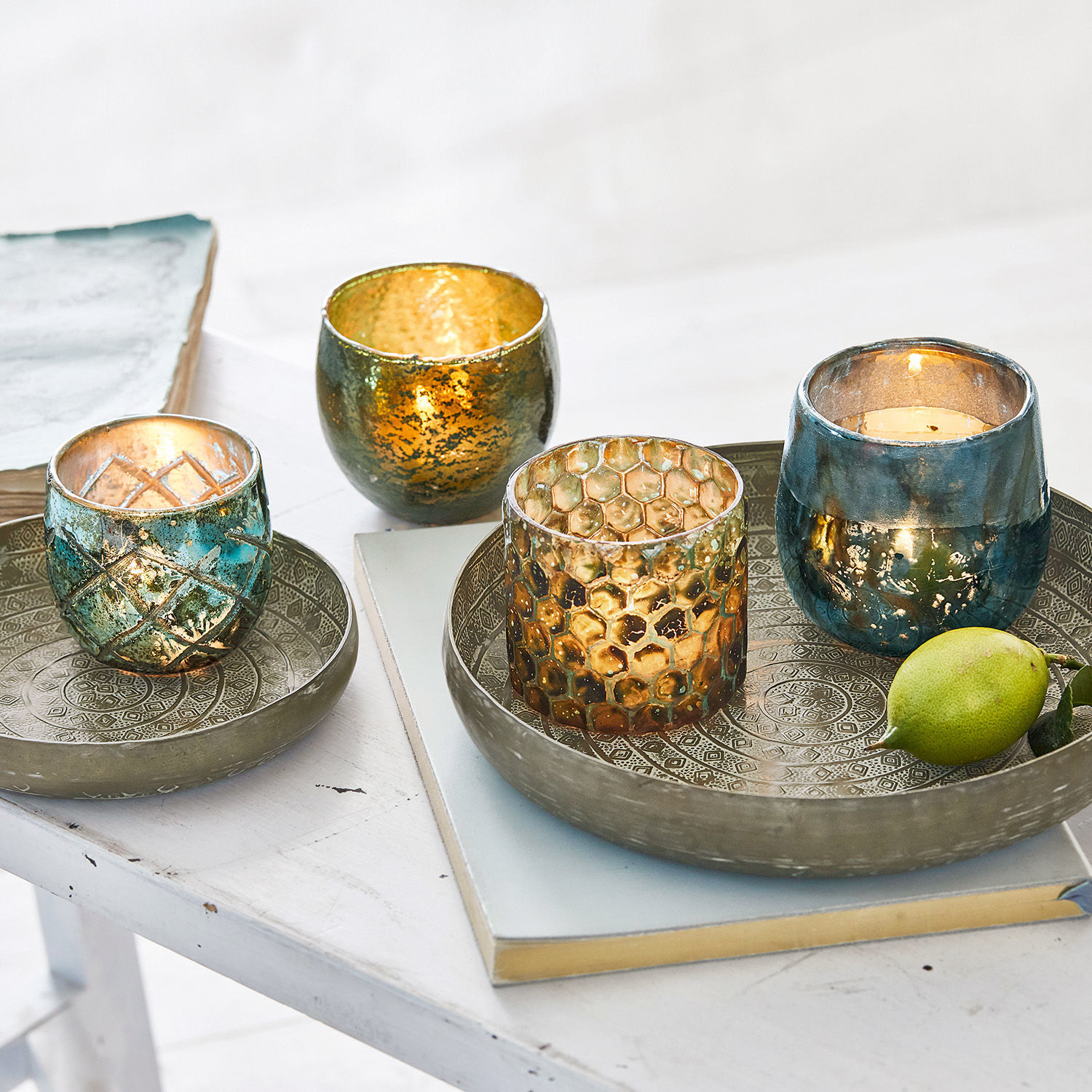 Set glazen theelichthouders in goudblauwe tinten op metalen schalen op een witte tafel