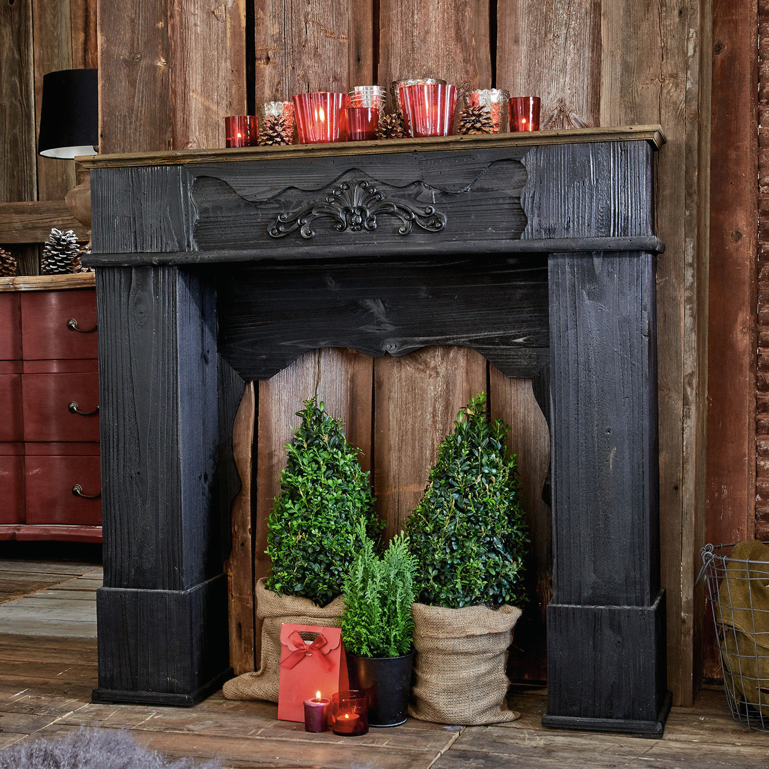 Haardschouw Margaux Zwarte houten decoratieve open haard met groene planten en rode kaarsen in rustieke kamer