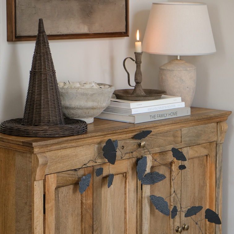 Houten dressoir met zwarte bladvormige slinger, lamp en kandelaar op boeken in woonkamer