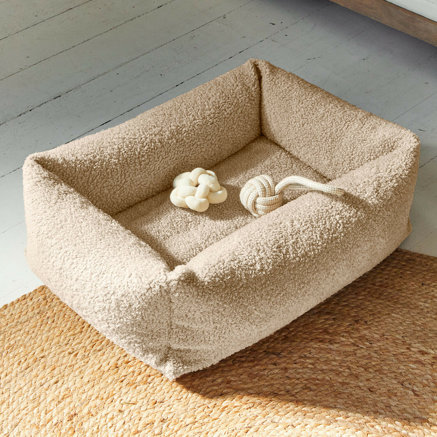 Beige hondenmand van zacht materiaal met twee beige knoop speeltjes op een vloer met gevlochten tapijt