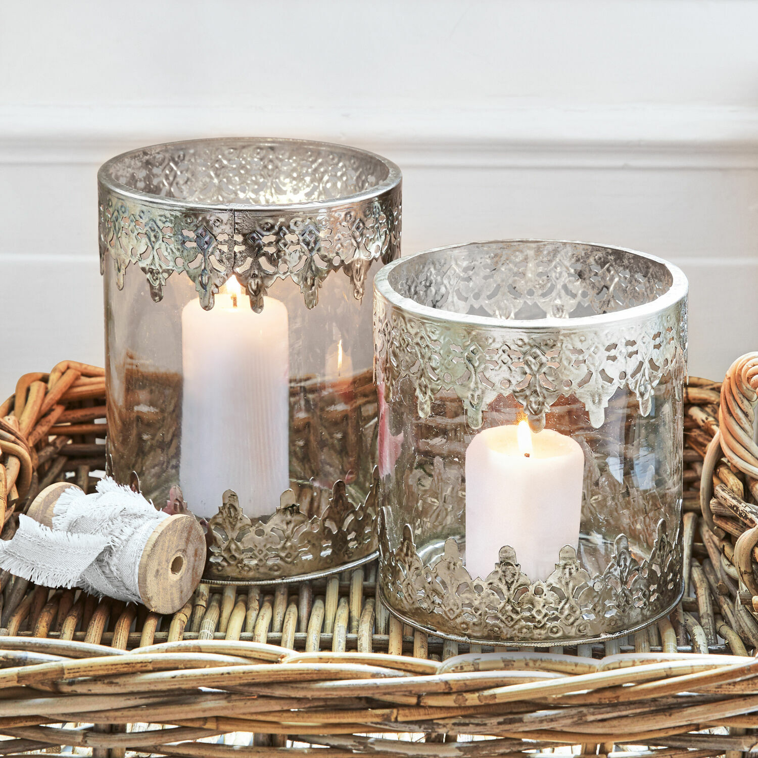 Twee glazen windlichten met zilveren metalen kanten decoratie op een rieten dienblad