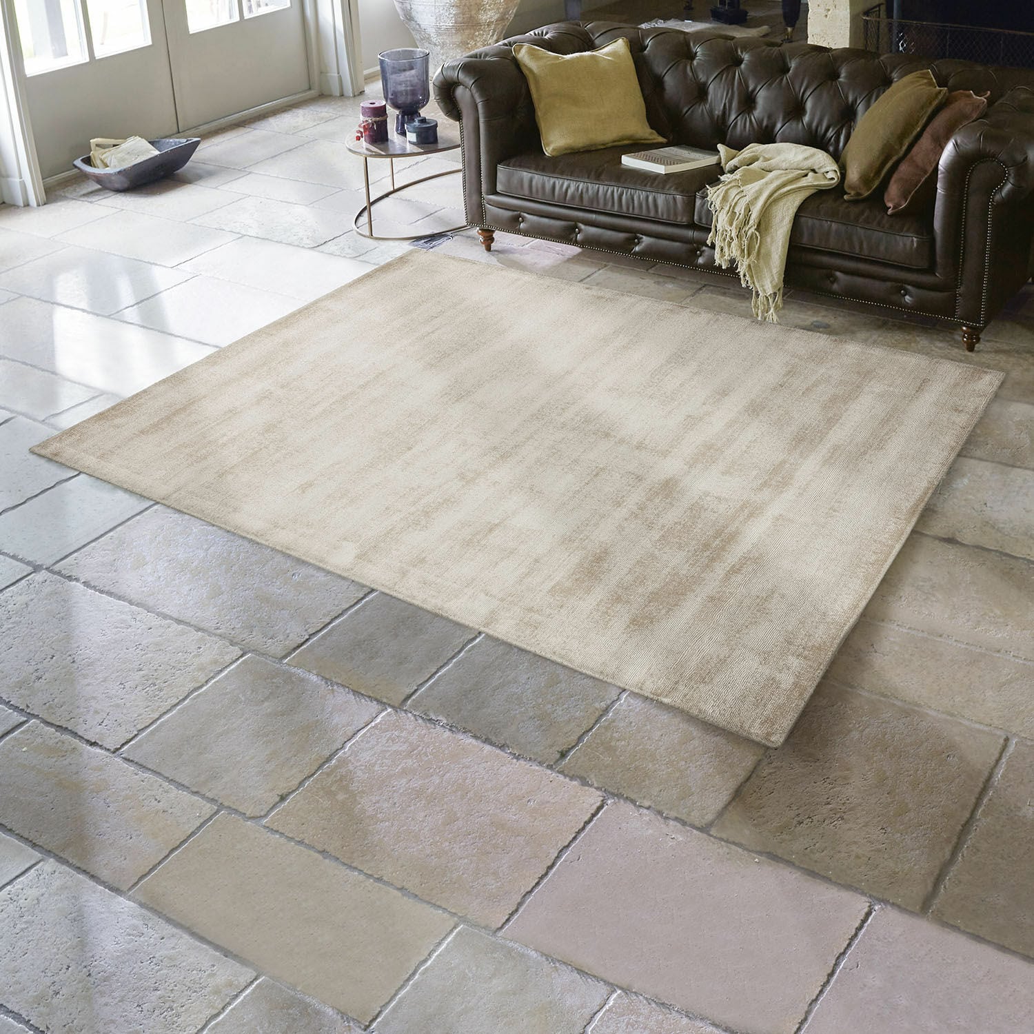 Vloerkleed Mariselle Beige vloerkleed van zacht materiaal op stenen vloer in woonkamer met bruine leren bank