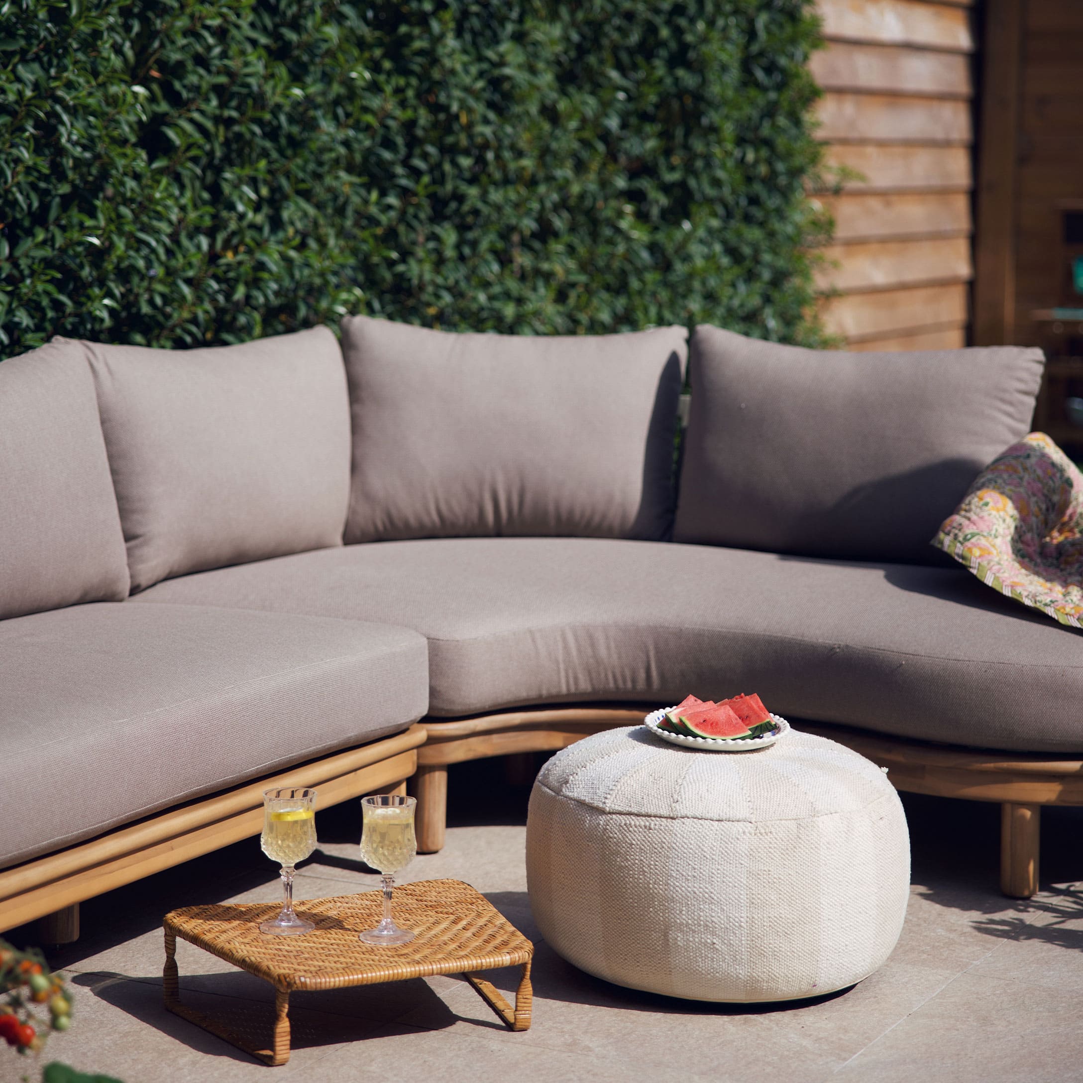 Beige bank met kussens op houten frame en witte poef op terras met zomerse decoratie