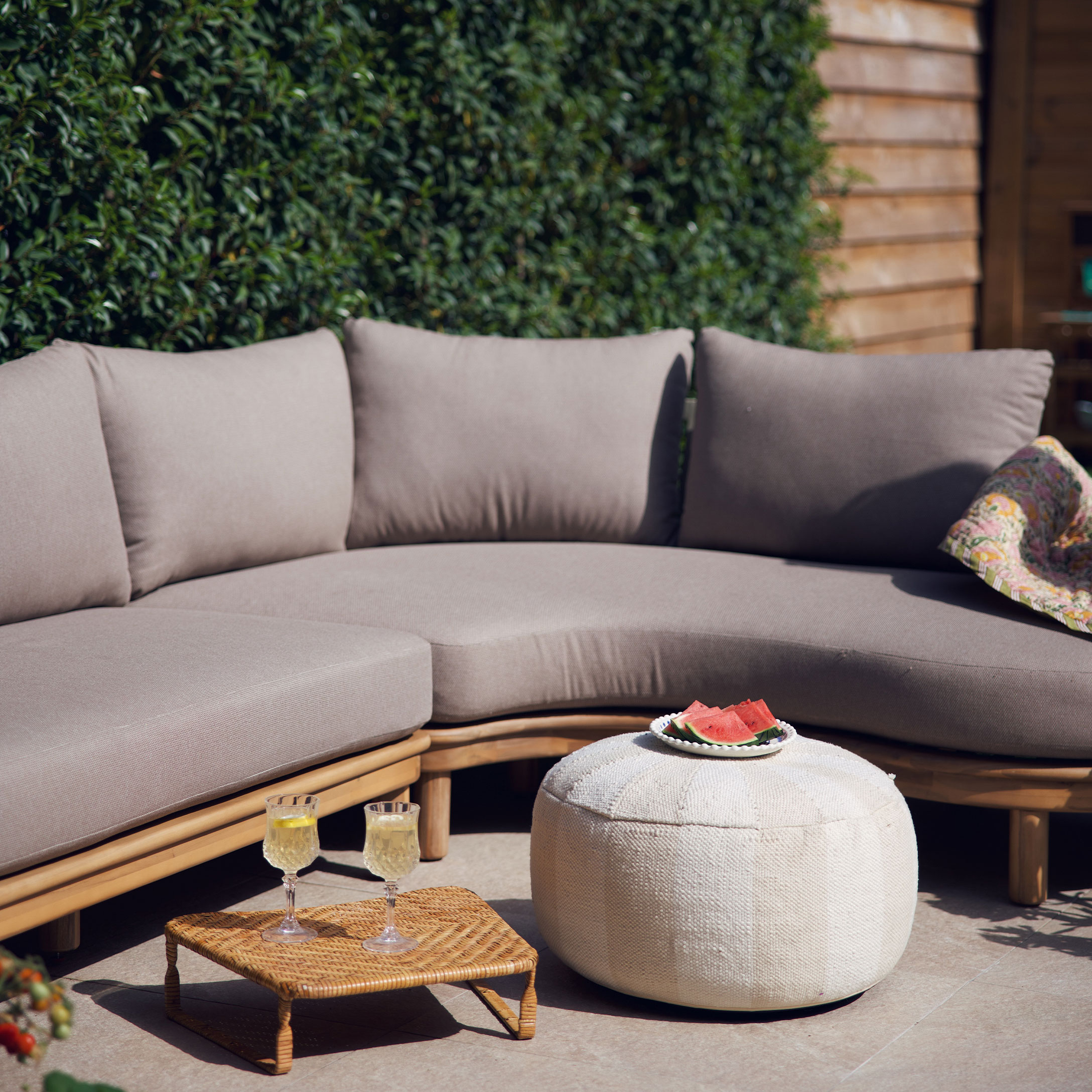 Beige bank met kussens op houten frame en witte poef op terras met zomerse decoratie