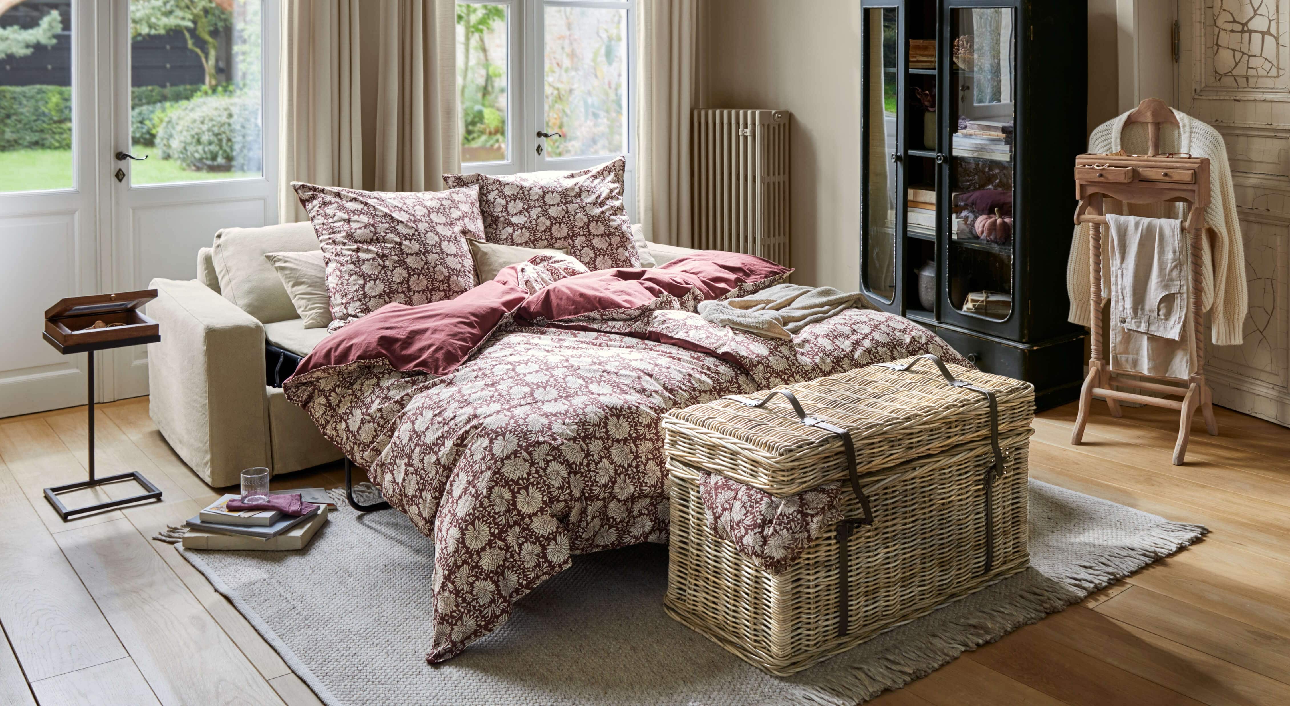 Uitschuifbare bank met beige bekleding en bordeaux bloemenbeddegoed in lichte woonkamer met houten vloer