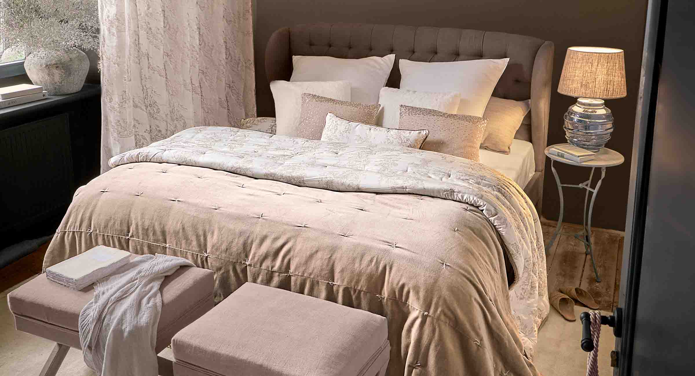 Gezellige slaapkamer met beige dekbed en kussens op gestoffeerd bed, lamp op nachtkastje