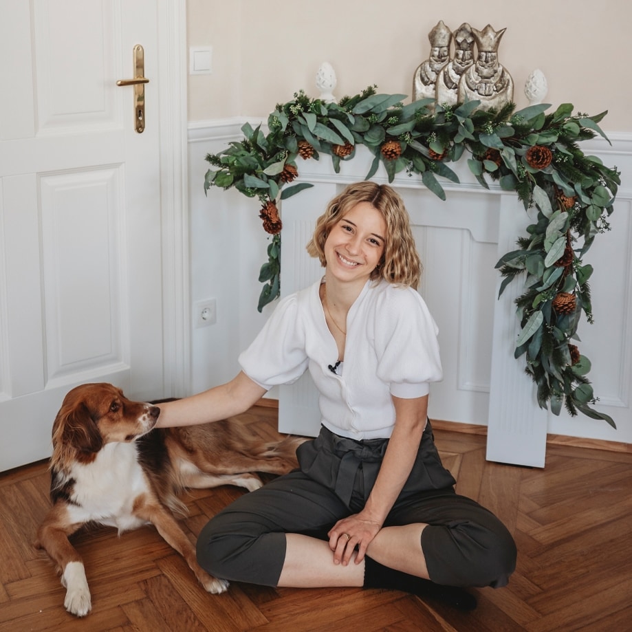 Jonge vrouw in witte trui en donkere broek zittend op houten vloer met hond bij kerstdecoratie van groene bladeren en dennenappels