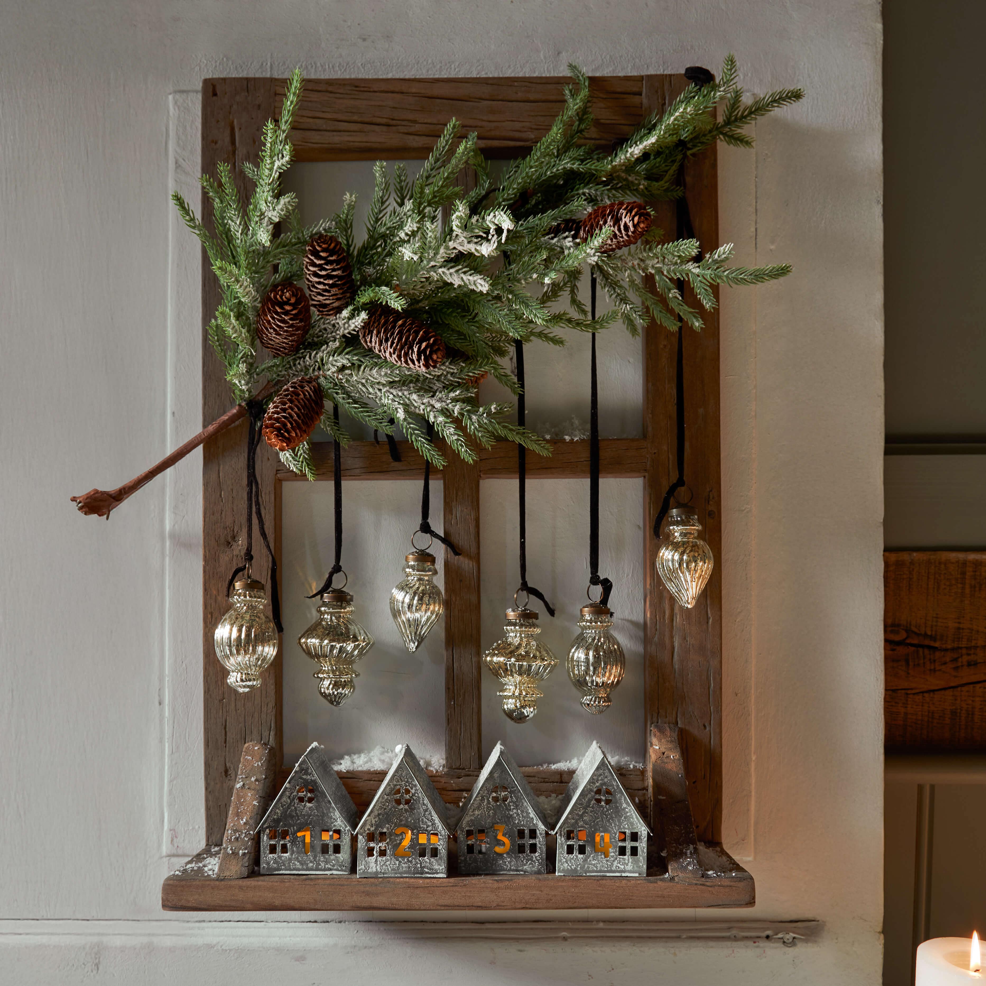 Houten lijst met genummerde metalen huisjes en hangende glazen ornamenten, kerstdecoratie met dennenappels en groene takken