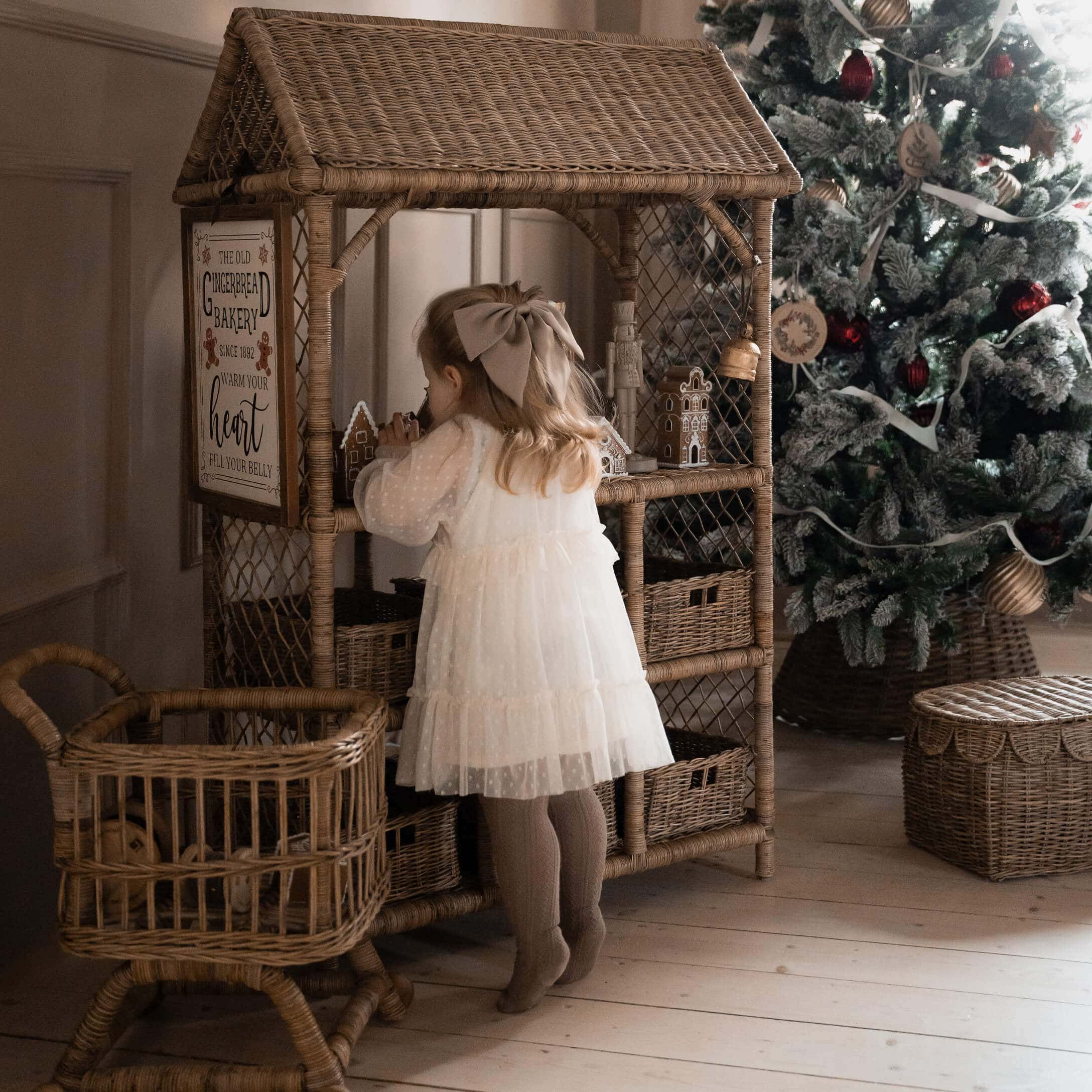 Kind in witte jurk bij rotan kast met peperkoekhuisjes naast versierde kerstboom en rotan manden