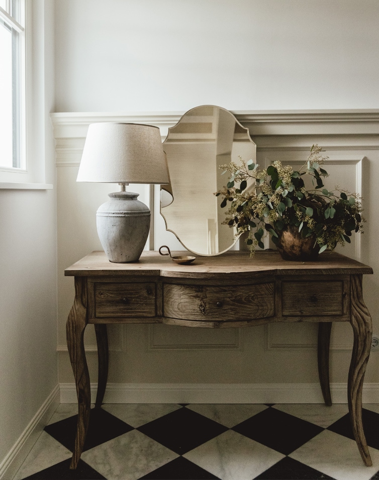 Houten bureau met spiegel, lamp en plantenversiering op zwart-witte vloer in lichte kamer