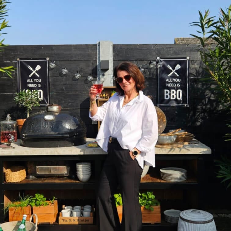 Vrouw in wit overhemd en zwarte broek bij buitenbarbecue met BBQ-bord tegen zwarte muur