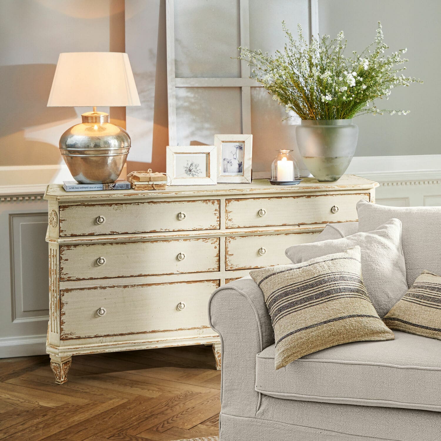 Ladekast Commeren Crèmekleurige houten vintage kast met lamp, fotolijsten en decoraties in woonkamer met beige bank