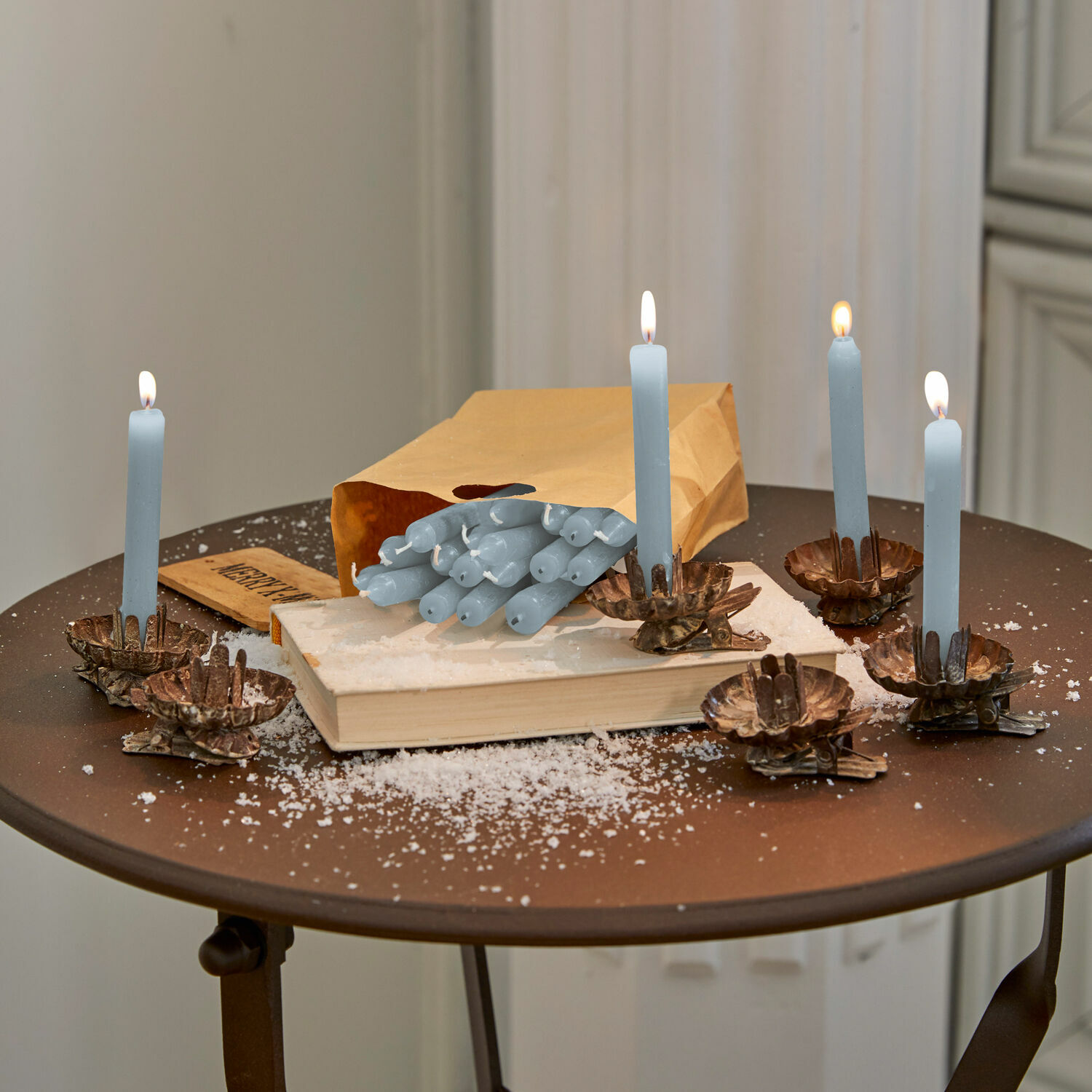Kaars set van 20 Noufa Blauwe kaarsen met messing kandelaren op bruine tafel met boek en papieren zak