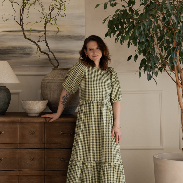 Vrouw in groene geruite jurk leunt op houten ladekast in gezellige kamer met planten