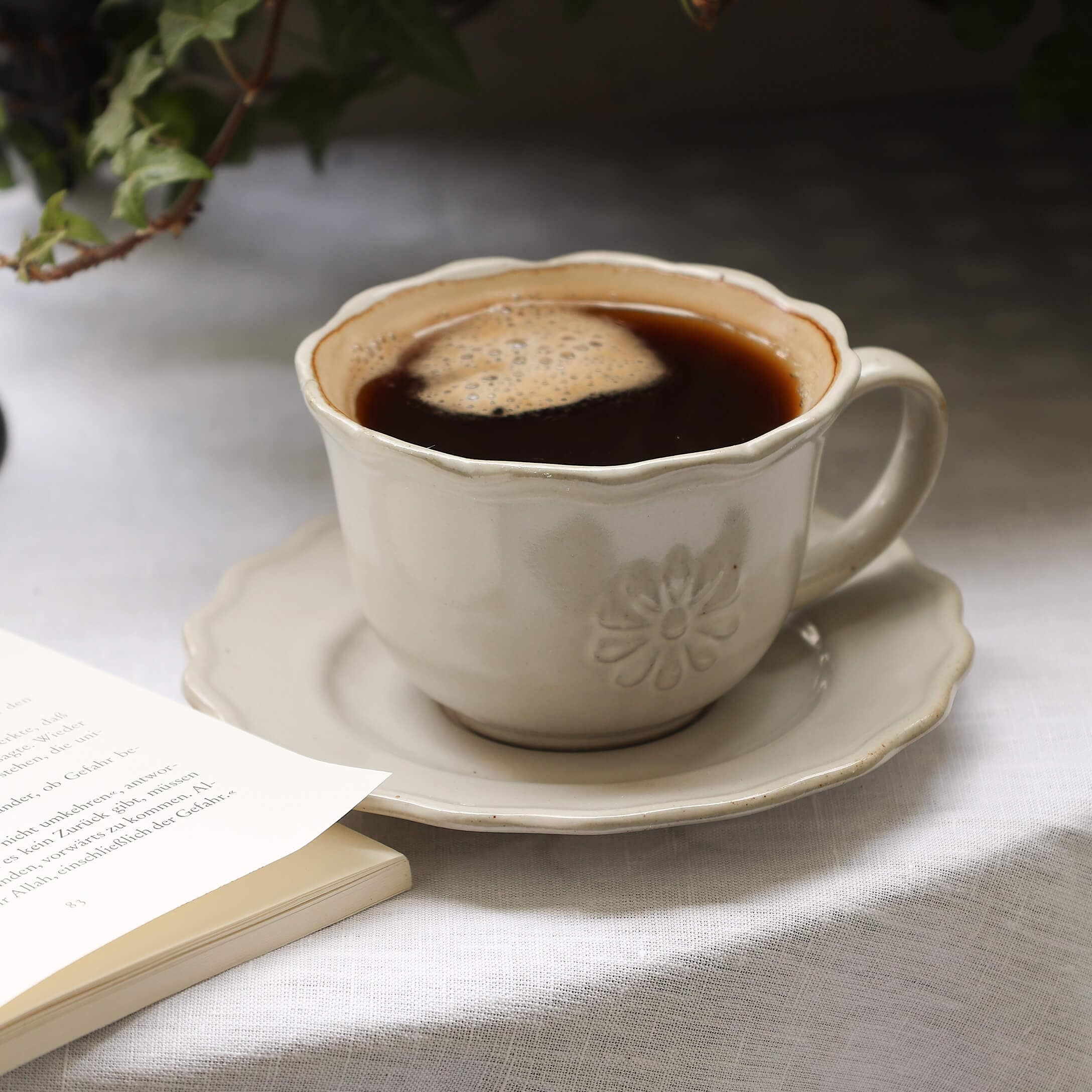 Beige keramische koffiekop met koffie op schotel op wit tafelkleed naast open boek