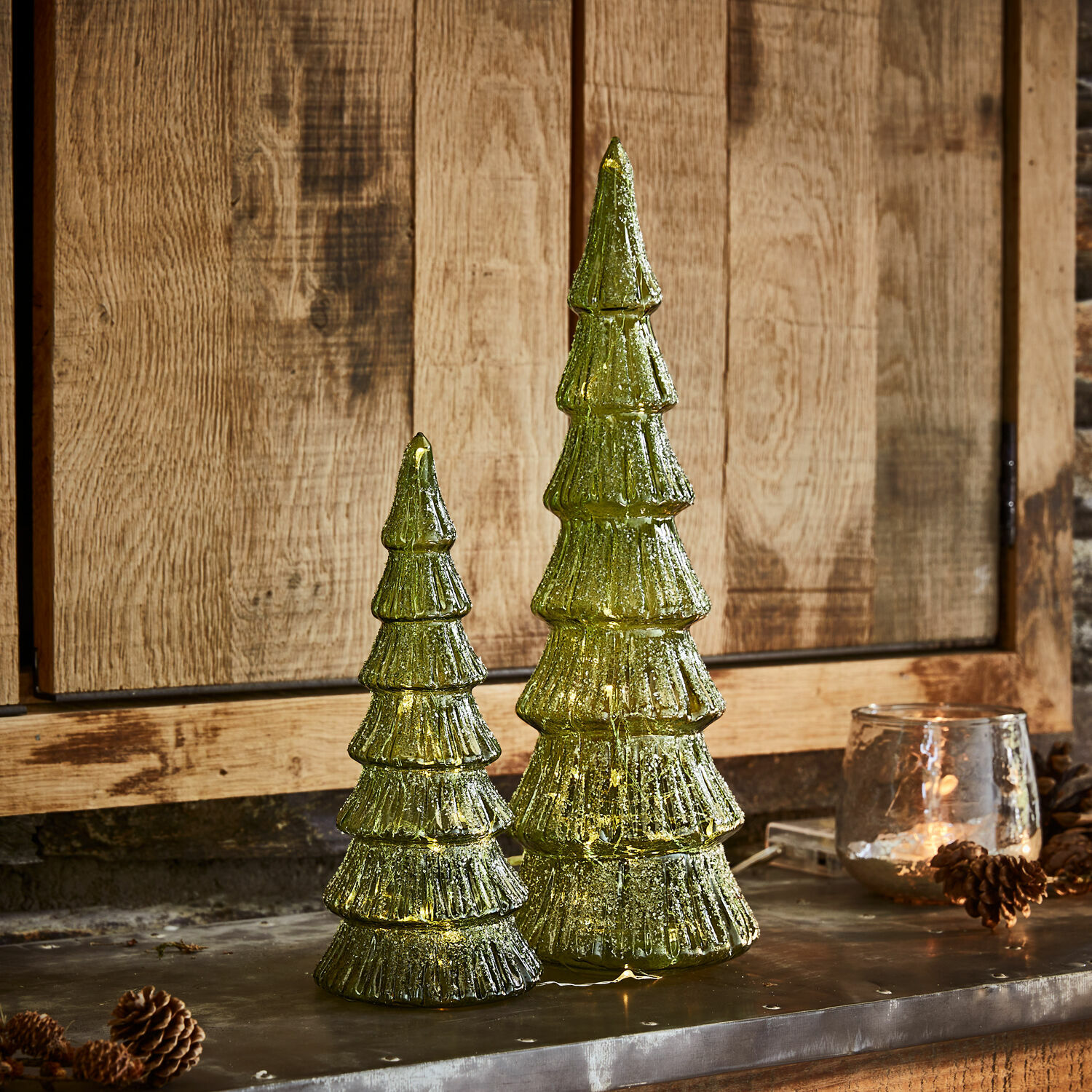 Ledkerstboom set van 2 Merrton Twee groene glazen kerstboomdecoraties met LED-verlichting tegen houten achtergrond, feestelijke versiering