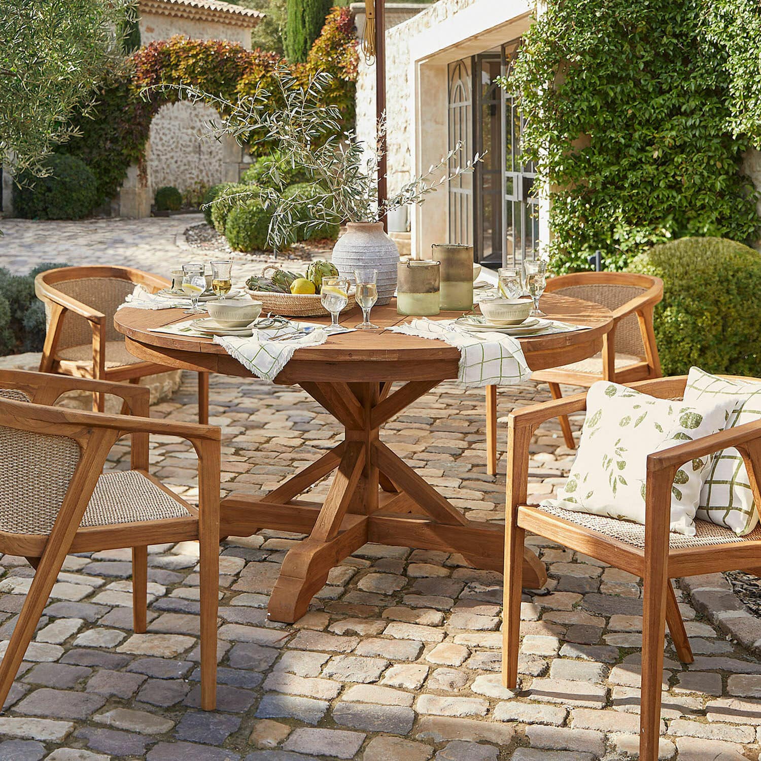 Tafel Wendreau Ronde teakhouten tafel met stoelen op een geplaveid terras, gedekt met servies en decoratie