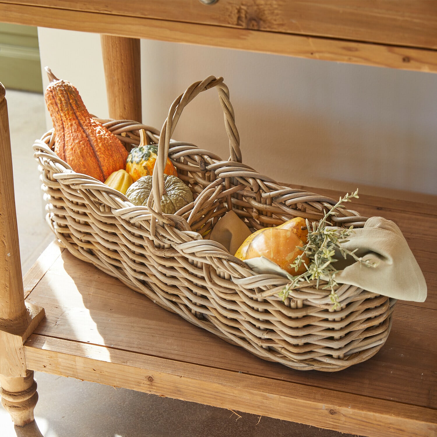 Mand Allegretta Gevlochten rieten mand met handvat gevuld met decoratieve pompoenen en groene takken op houten plank