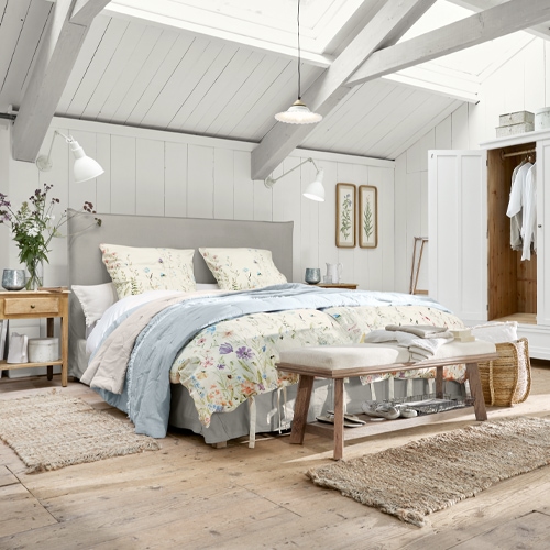 Gezellige slaapkamer met lichtgrijs gestoffeerd bed, bloemrijk beddengoed, natuurlijk hout en vloerkleden