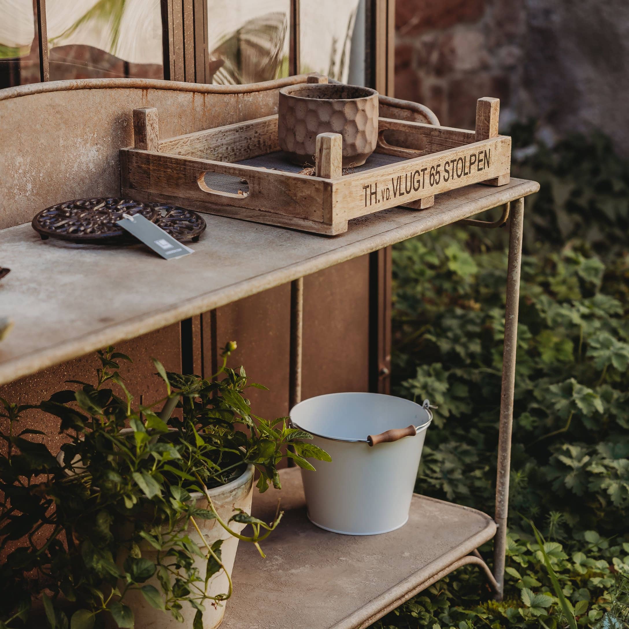 Houten dienblad op metalen tafel met bloempot en witte emmer in tuin als decoratie