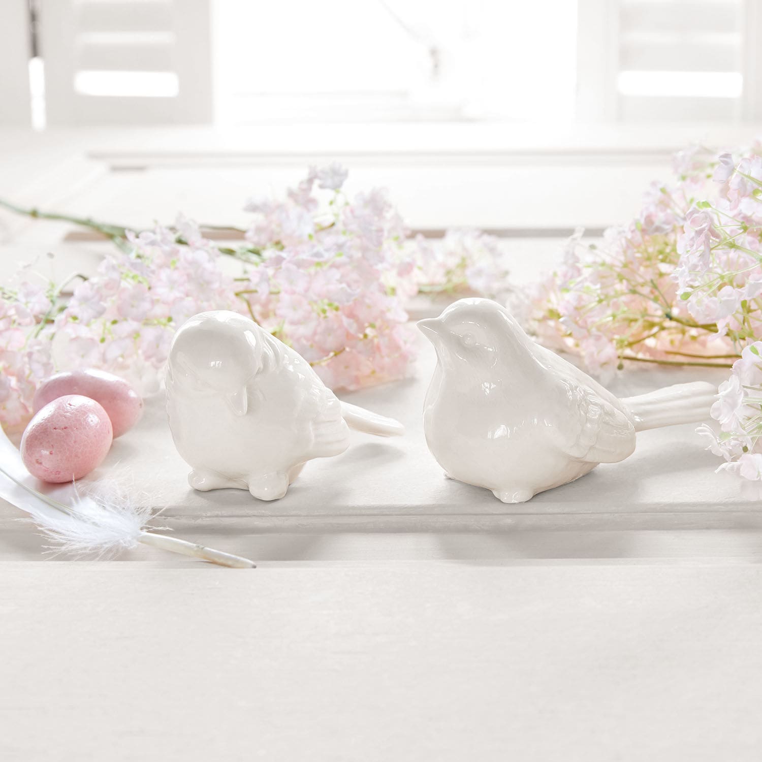 Twee witte keramische vogelbeeldjes op lichte achtergrond met roze bloemen en decoratieve eieren