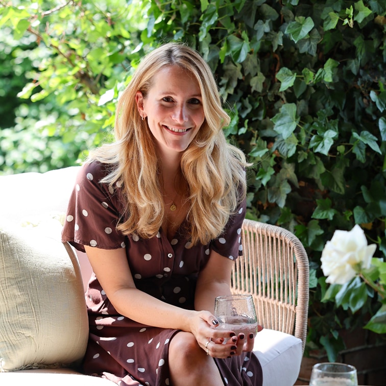 Vrouw in bruin polkadotjurk zittend op rotan stoel met glas in hand in tuinomgeving