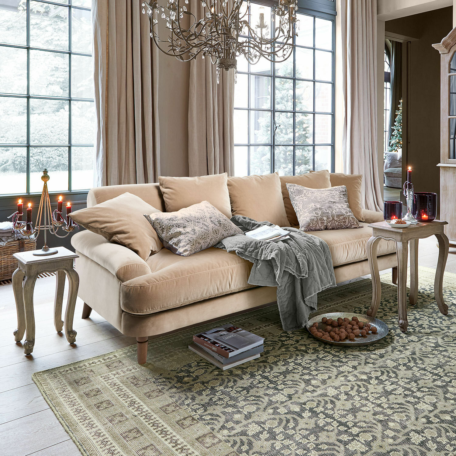 Bank Rivermere Beige fluwelen bank met kussens en grijze deken in lichte woonkamer met tapijt en kaarsen