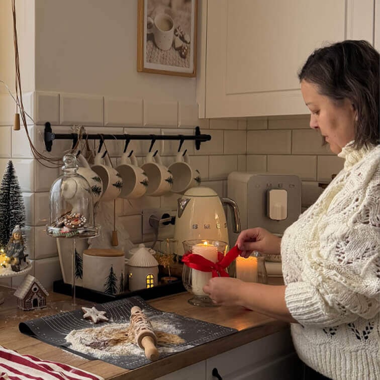Vrouw in witte trui versiert kaars met rood lint in keuken met houten werkblad en kerstdecoratie