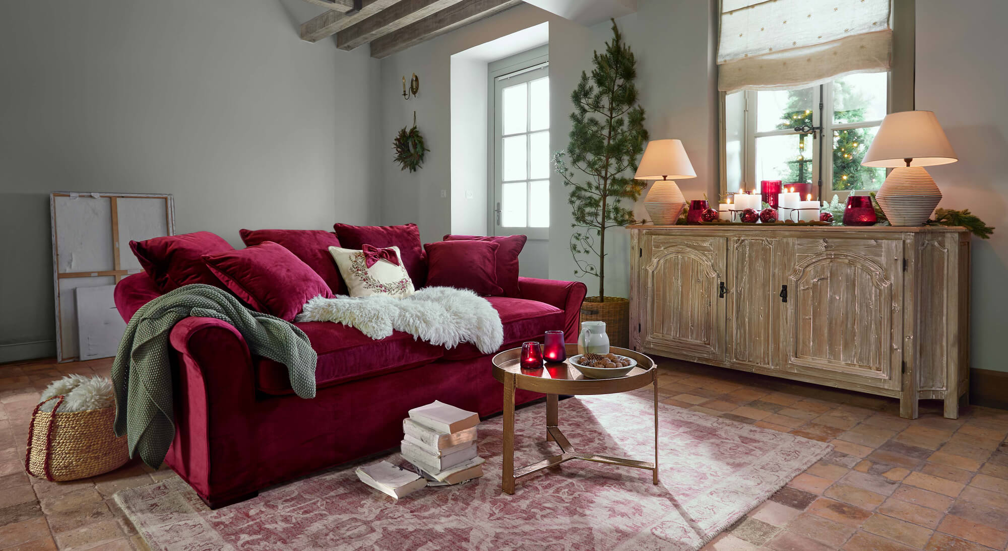 Rode fluwelen bank met kussens en groene deken in gezellige woonkamer met houten dressoir en kerstdecoratie