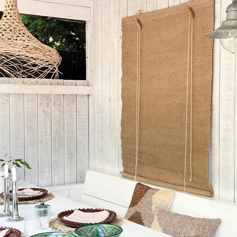Witte houten wand met natuurlijke jute rolgordijn en gedekte tafel met kussens in boho stijl