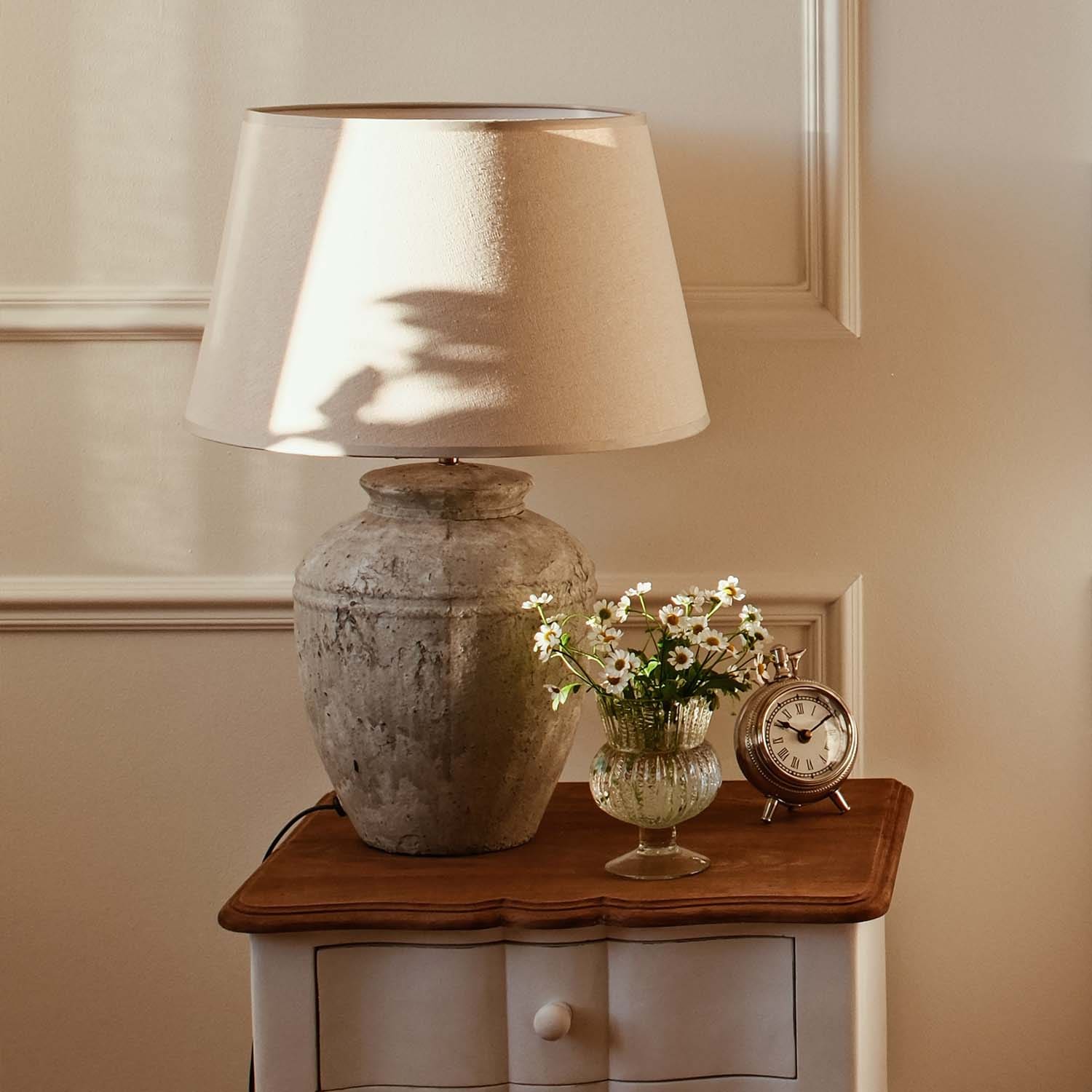 Tafellamp met beige kap en vintage keramische voet op houten tafel met bloemen en klok