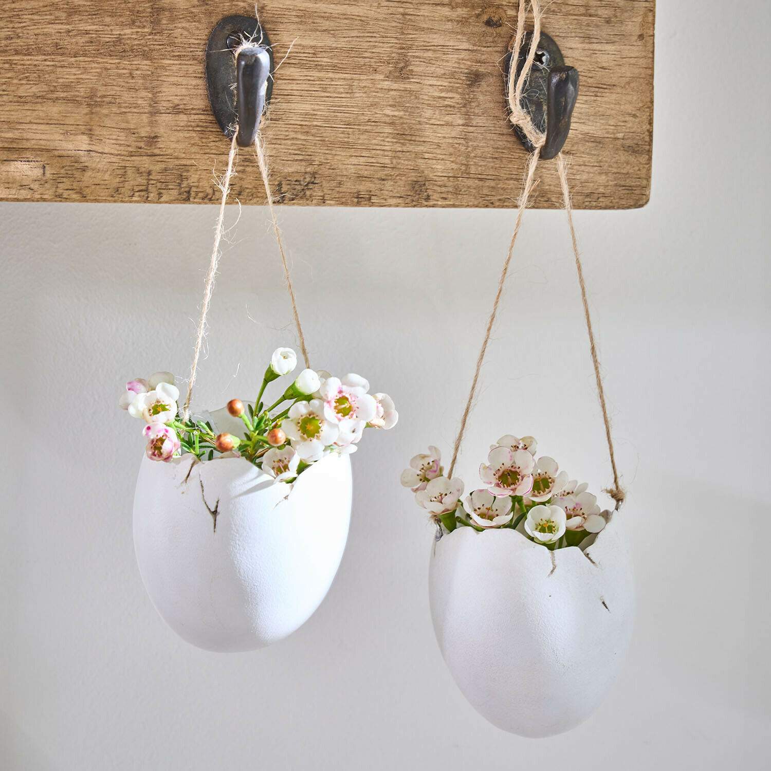 Hangvaas set van 2 Nalena Twee witte keramische bloempotten in eivorm met bloemen, hangend aan touwen aan een houten haak