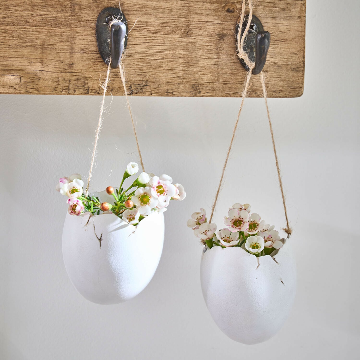 Hangvaas set van 2 Nalena Twee witte keramische bloempotten in eivorm met bloemen, hangend aan touwen aan een houten haak